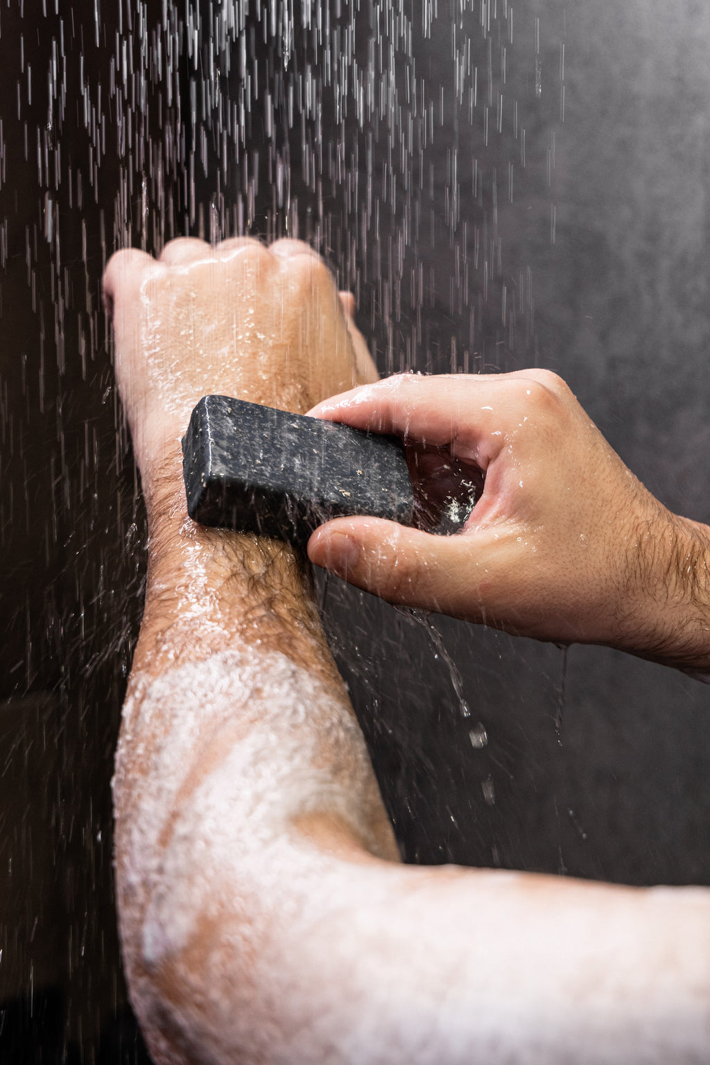 A close-up of a person's forearm and hand being washed with soap under running water, holding a dark rectangular bar of soap.