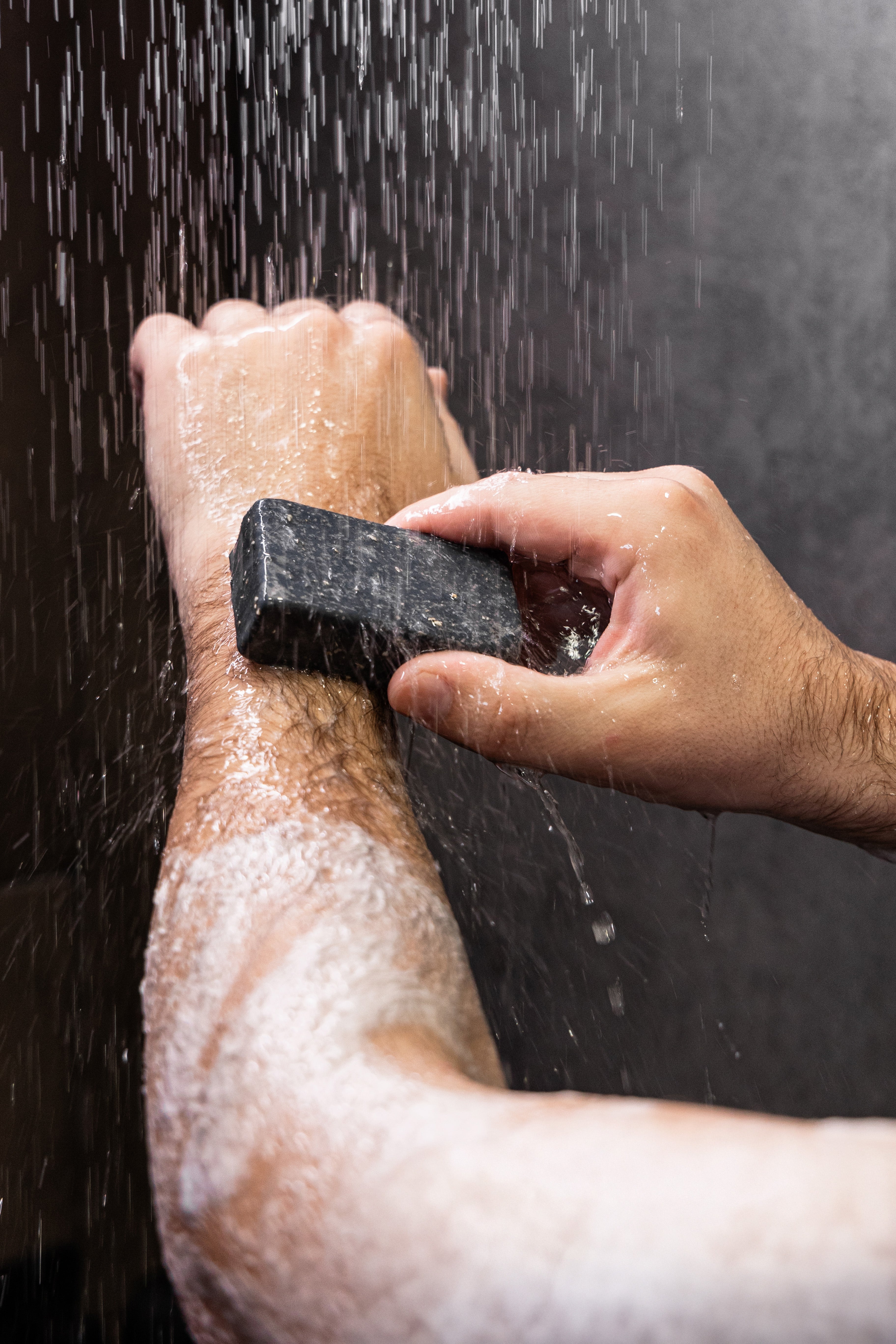 A close-up of a person's forearm and hand being washed with soap under running water, holding a dark rectangular bar of soap.
